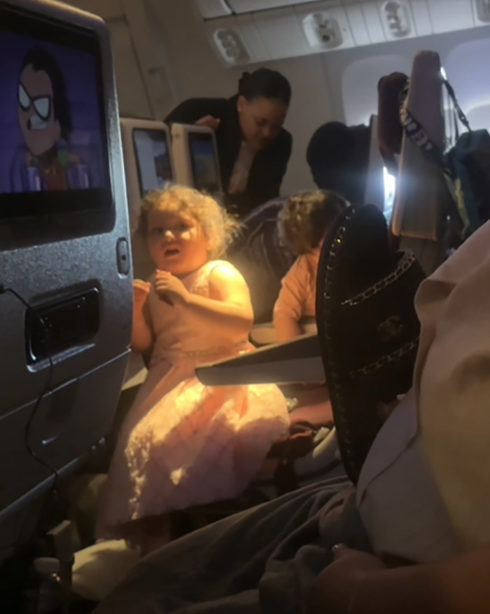 Child in pink dress on airplane, standing in aisle with person nearby; in-flight screen visible.