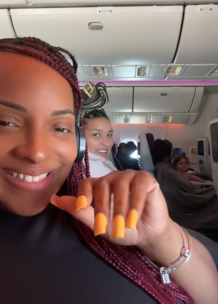 Passengers on a flight, one smiling woman in focus, others seated, overhead compartments visible.