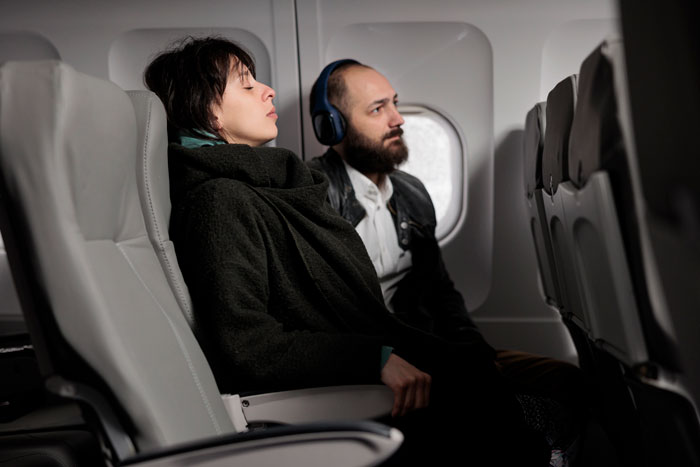 Two passengers on a plane, one occupying a window seat with headphones, the other seated beside, both looking forward. Two passengers on a plane, one occupying a window seat with headphones, the other seated beside, both looking forward.