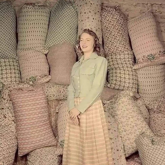 A woman in vintage attire stands smiling in front of a stack of patterned sacks, capturing a forbidden story theme.