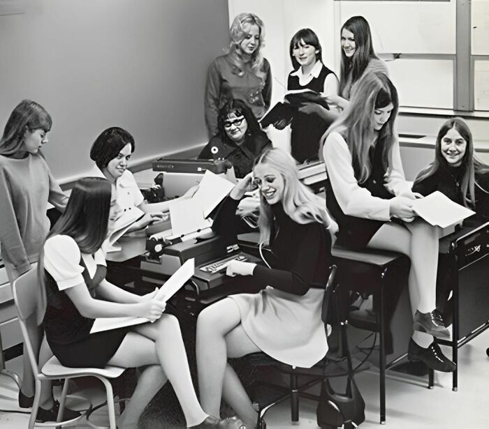 Group of women in a 1970s classroom setting, engaging with typewriters and papers, reflecting forbidden stories theme.