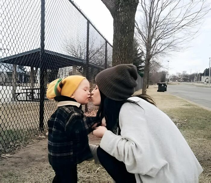 Mother and child share a sweet moment beside a park fence in their winter attire, illustrating a touching forbidden story.