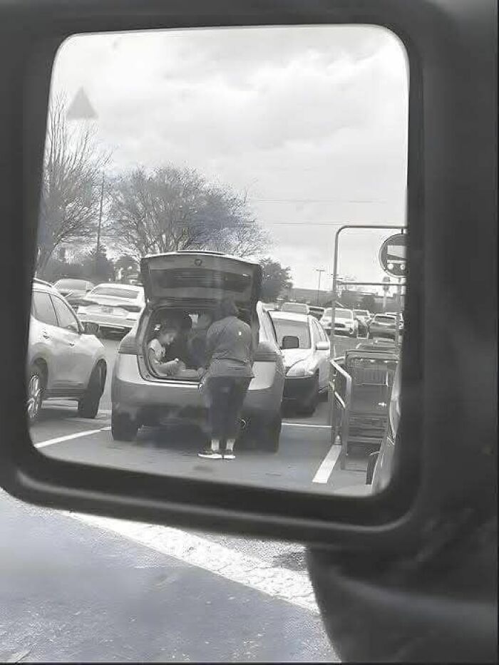 Person loads a car trunk in a parking lot, captured through a side mirror, depicting an urban scene.