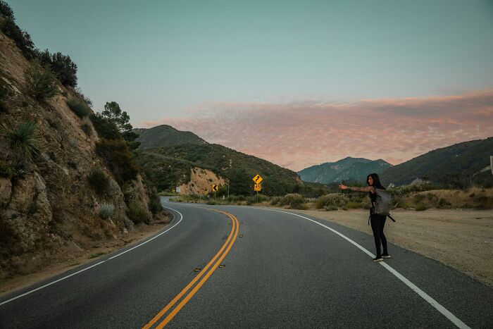 Person hitchhiking on a scenic USA road at sunset, highlighting culture shock.
