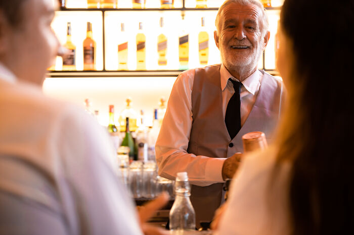 Elderly bartender smiling while preparing drinks, highlighting culture shocks in USA nightlife.