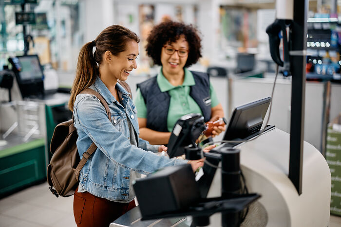 A woman experiences a culture shock in a USA supermarket while interacting with a smiling cashier at the checkout.