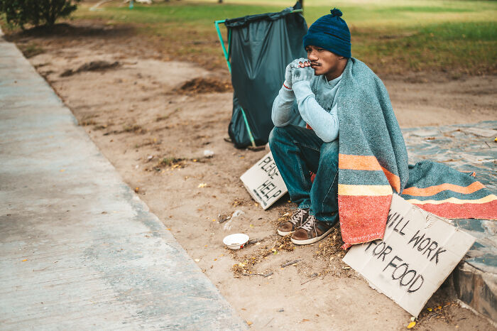 A person experiencing culture shock in the USA, sitting outside wrapped in a blanket, with signs seeking help.