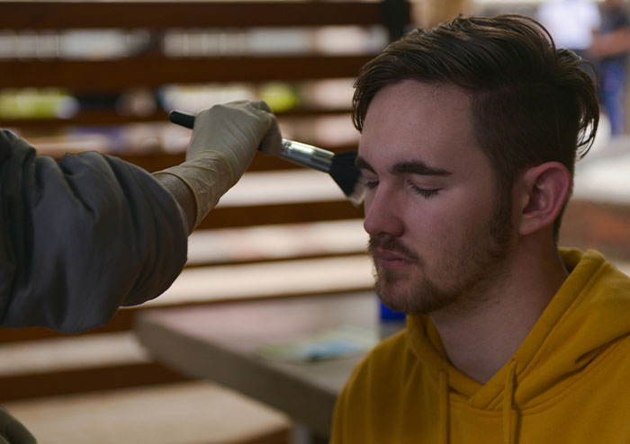 Man in a yellow hoodie getting makeup applied, related to a celebrity's online confession about feeling tired.