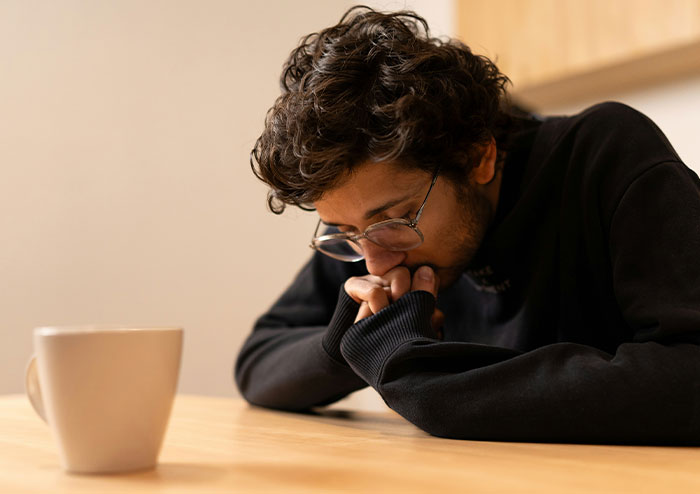 Individual in glasses sitting at a table, appearing contemplative and tired, with a mug nearby.