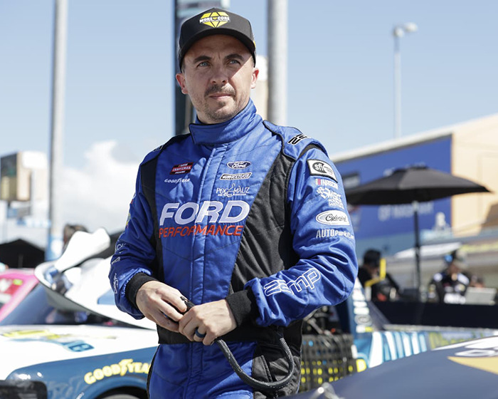 Frankie Muniz in a Ford racing suit, standing confidently at a car event, showcasing "Malcolm in the Middle" nostalgia.