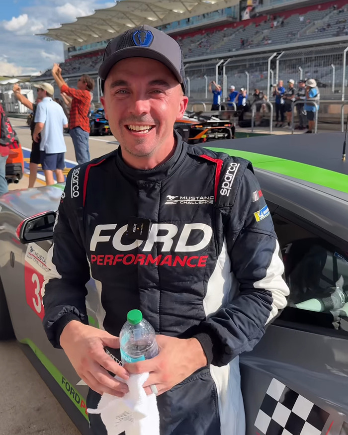 Man in racing suit and cap holding water bottle at a racetrack, smiling after a 'Malcolm In The Middle' reunion.