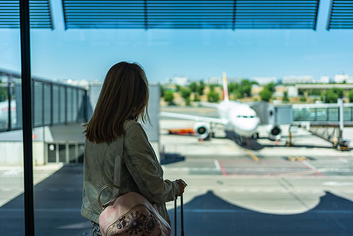 Woman at airport window with suitcase, looking at plane on runway.