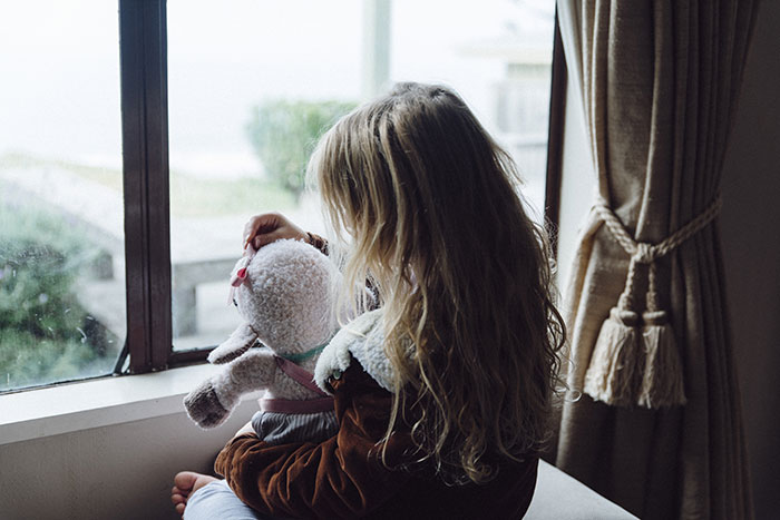 Young girl holding a stuffed toy, gazing out of a window, symbolizing waiting and uncertainty.