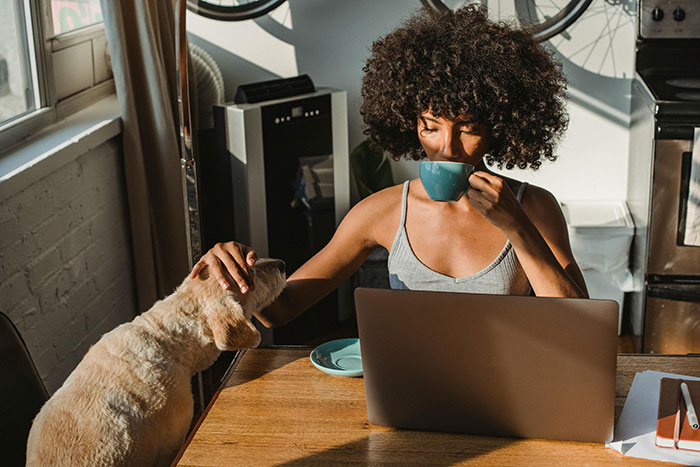 Woman drinking coffee at a table, petting a dog, with a laptop, reflecting casual birthday planning and diet challenges.