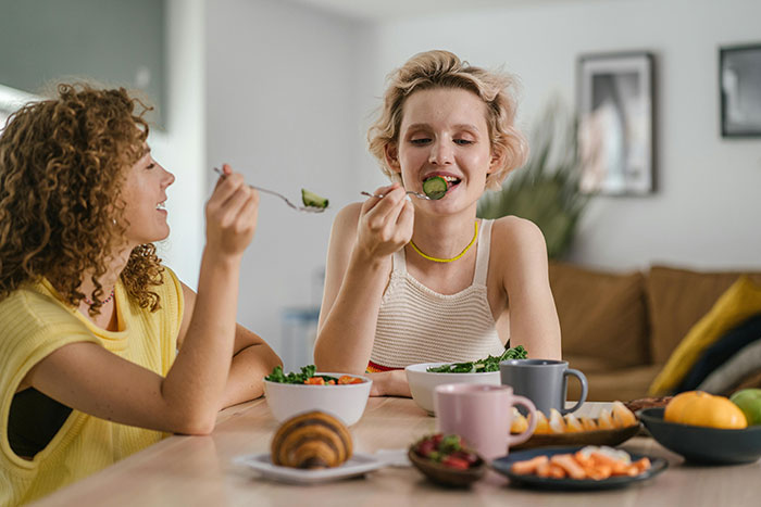Two friends eating at a table, enjoying a meal with varied dishes, smiling and discussing birthday plans.
