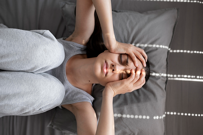 Woman in gray loungewear lying on a bed, holding her head, experiencing anxiety before a wedding.