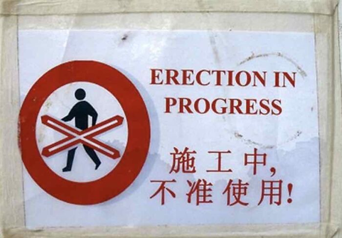 Warning sign humorously incorrect, reading "Erection in Progress" with crossed-out pedestrian symbol.