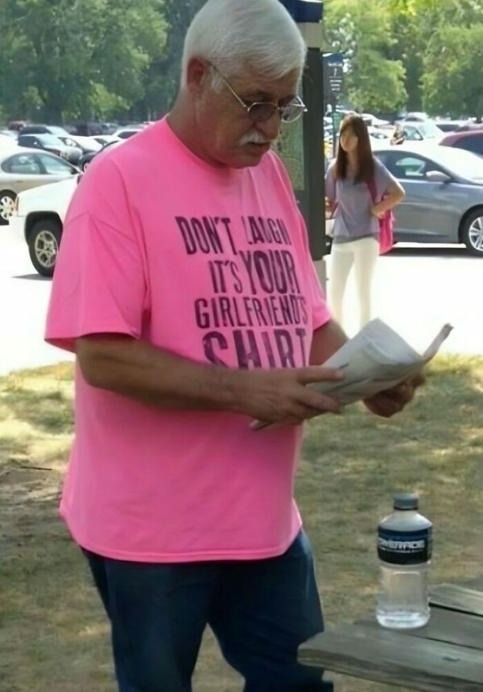 Hombre mayor con camiseta rosa divertida leyendo periódico en parque con coches de fondo, camisetas ridículas.