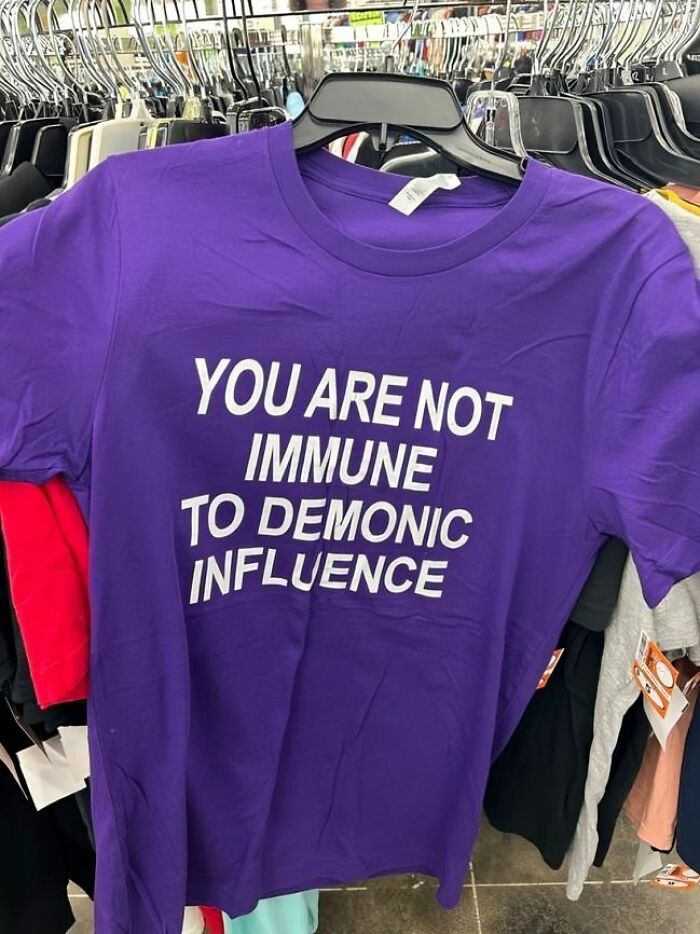 Camiseta divertida y ridícula color morado con texto en inglés, colgada en tienda entre otras prendas variadas.