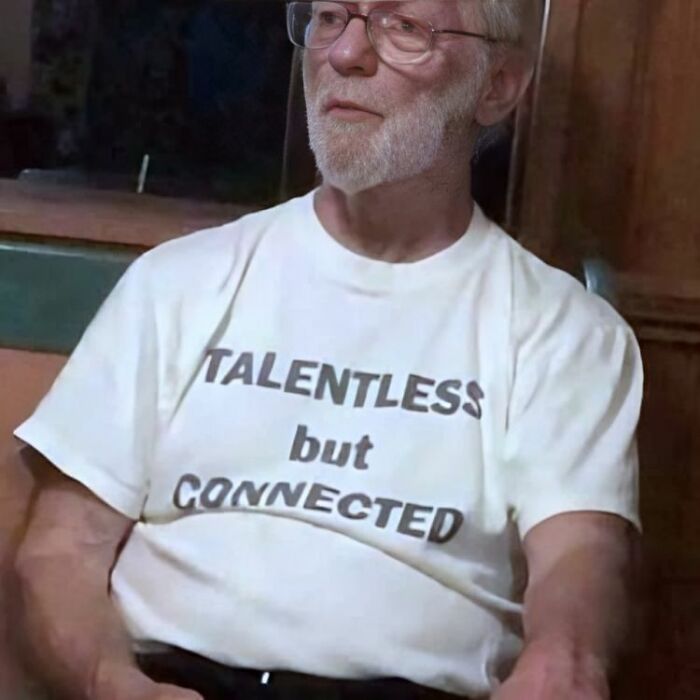 Hombre mayor con camiseta divertida y ridícula con texto en inglés sentado en interior con fondo oscuro.