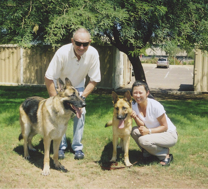 Man and woman with two German Shepherds on grass, smiling under a tree.