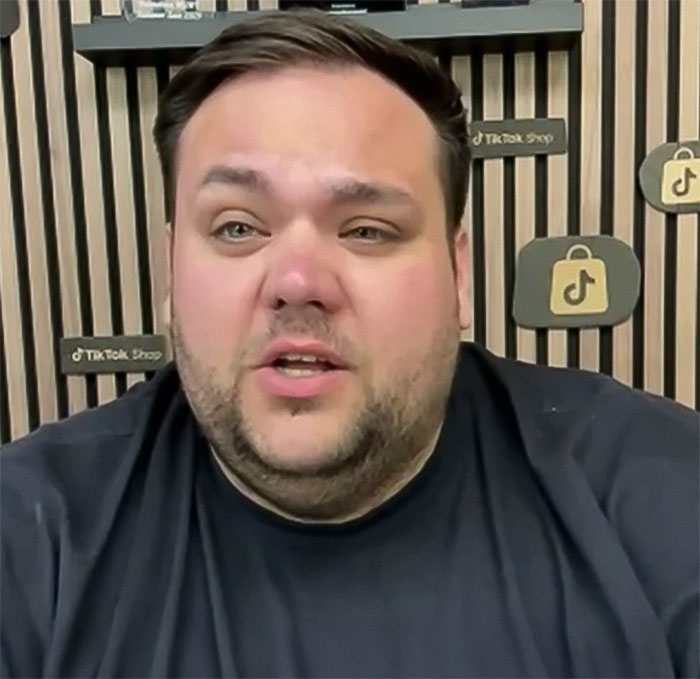 Plus-size dancer in black shirt speaks, wooden wall with TikTok logos in the background.
