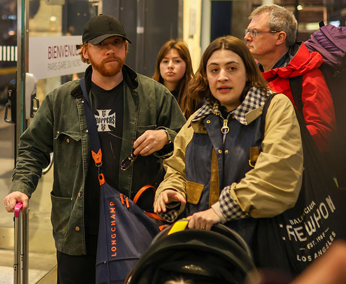 A man and a woman with luggage at a station, the man wearing a cap and jacket, the woman pushing a stroller.