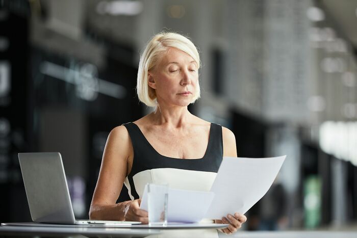 A woman in a black and white dress reads documents, skeptical expression, sitting at a desk with a laptop during April 1st.