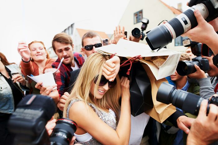 A woman with sunglasses surrounded by paparazzi, confidently shielding herself with shopping bags.