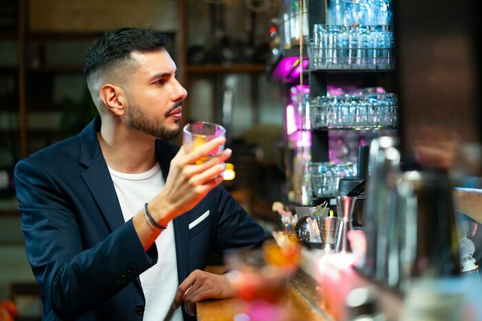 A man in a bar holding a drink, gazing towards the bartender, surrounded by glasses, experiencing something appalling.