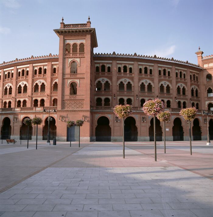 Historic arena with arches and towers in sunlight, surrounded by a plaza with trees.