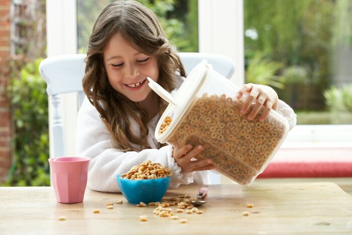 Girl pouring cereal from a large container into a bowl, with a pink cup beside her on the table.