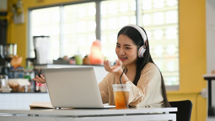 Person with headphones on a video call, smiling while gesturing at the laptop, with iced tea on the desk.