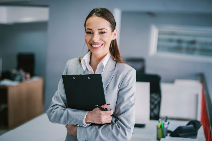 Woman in an office, smiling and holding a clipboard, representing American experiences shared internationally.