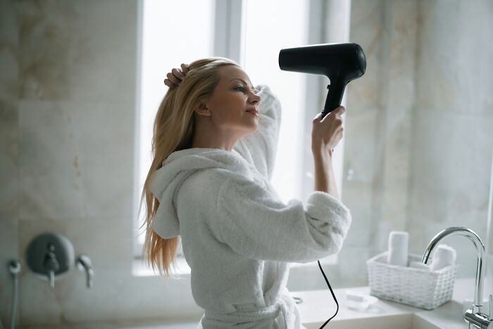 A woman drying her hair with a hairdryer in a European-style bathroom, wearing a white robe.