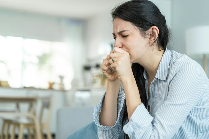 Woman in a light blue shirt sitting on a couch, looking thoughtful, with hands clasped near mouth.