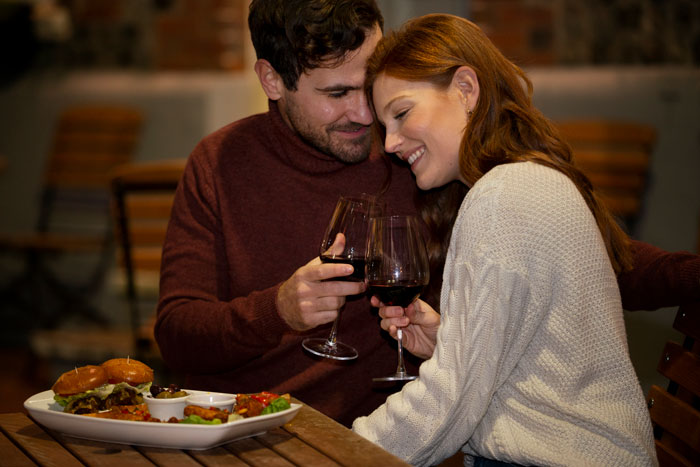 Couple on a cozy date night, smiling and clinking wine glasses at an outdoor restaurant.