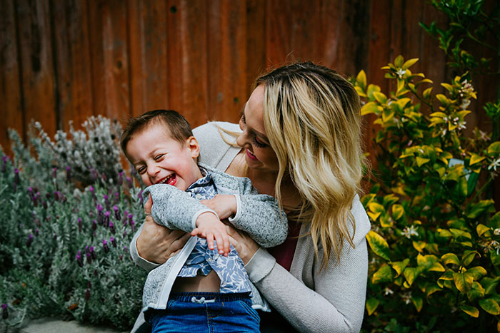 Mother and toddler son laughing together outside, surrounded by plants and a wooden fence.