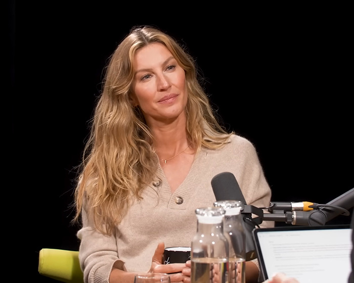 A person sits at a microphone, wearing a beige sweater, with studio equipment in the foreground.