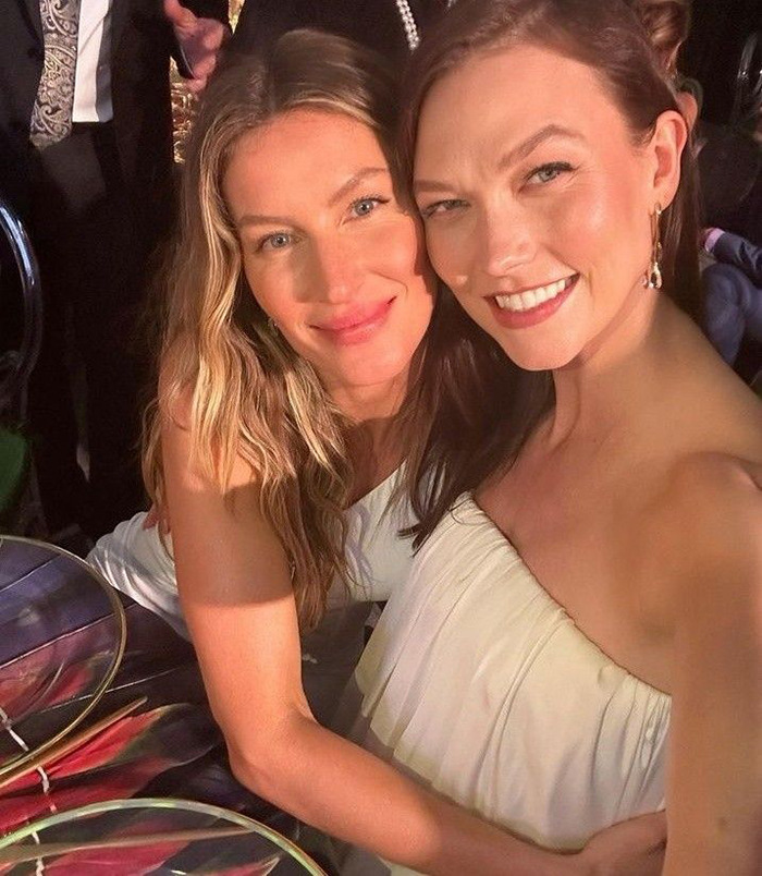 Two women smiling, posing together in a warmly lit setting, embracing while seated at a table.