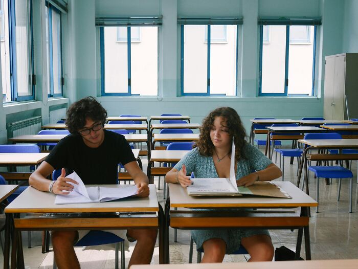 Two young people sitting at separate desks in a bright classroom, appearing to have a playful relationship argument.