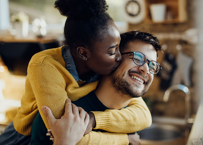 Woman hugging and kissing a smiling man in a cozy kitchen setting, showcasing partner affection.
