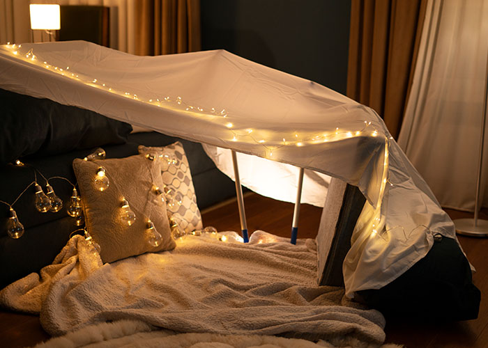 Cozy blanket fort in a dimly lit room, draped with string lights, creating a warm and inviting atmosphere.