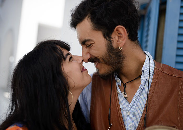 A happy couple smiling and touching noses, wearing casual outfits, standing close together in an outdoor setting.