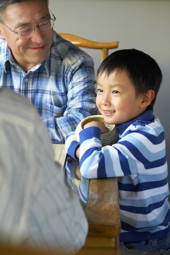 Child in a striped sweater smiling at a table, with an adult nearby, showcasing children's comedic nature.