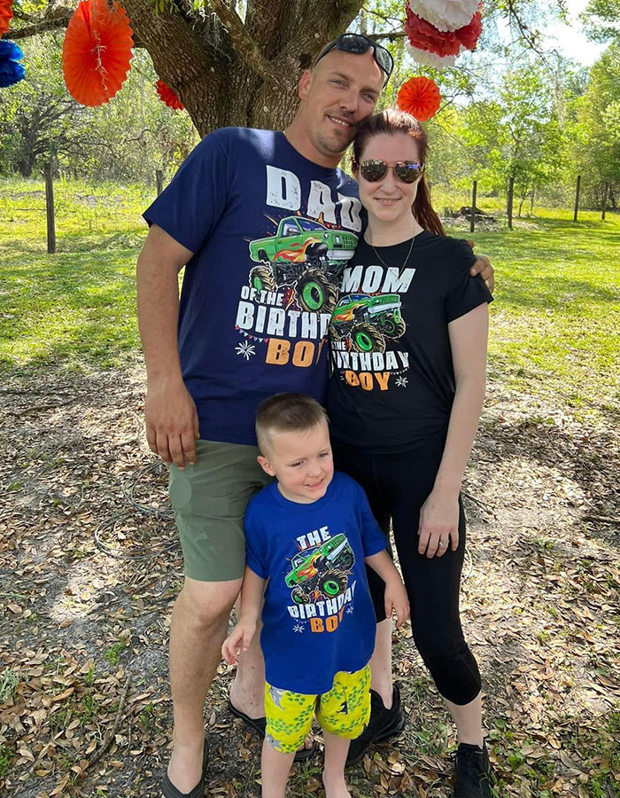 A family of three posing outdoors in coordinated birthday shirts, under colorful decorations on a sunny day. A family of three posing outdoors in coordinated birthday shirts, under colorful decorations on a sunny day.