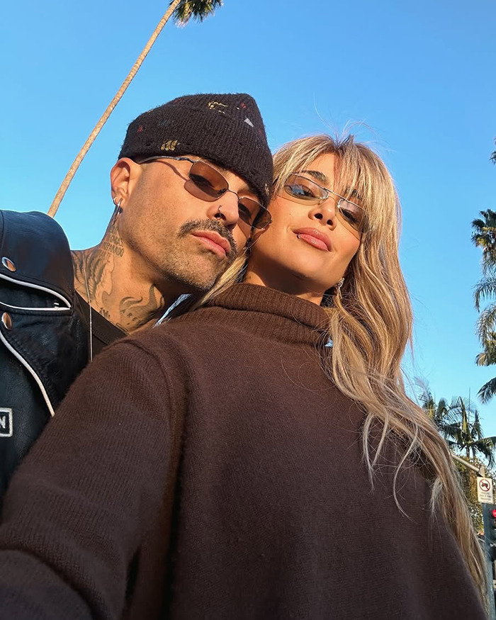 Two people in sunglasses under palm trees, blue sky, related to singer Greeicy's "husband stitch" revelation.