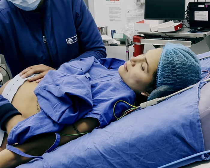 A woman in a hospital bed post-birth, attended by a doctor, wearing blue medical attire and cap.