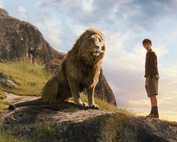 Scene from Narnia reboot with a lion and a boy on a rocky hill under a cloudy sky.