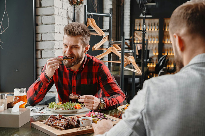 Man in a red flannel enjoying ribs at a restaurant, known for ordering more than friends during meals.